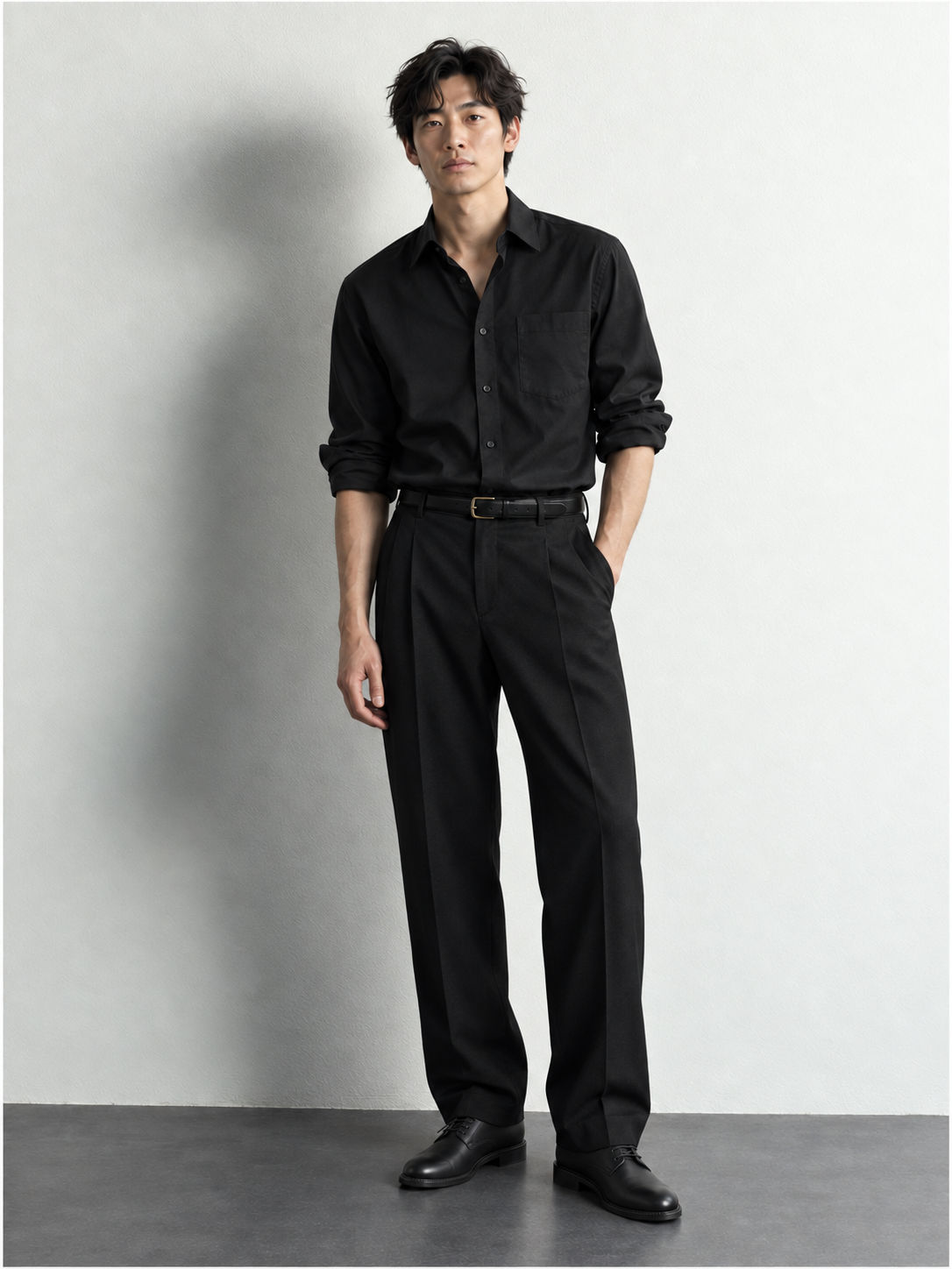 Full-length Maison 01 silhouette in a black shirt, black pleated trousers and black leather derby shoes.