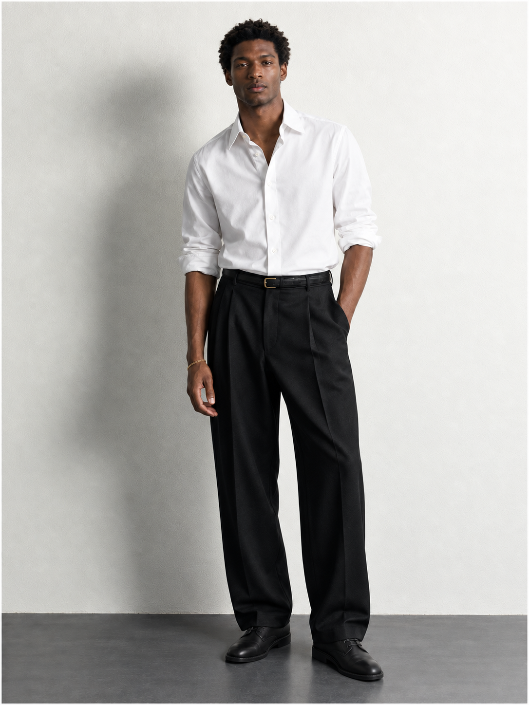 Full-length Maison 01 lookbook silhouette: a white shirt, black pleated trousers and black leather derby shoes against a pale wall.