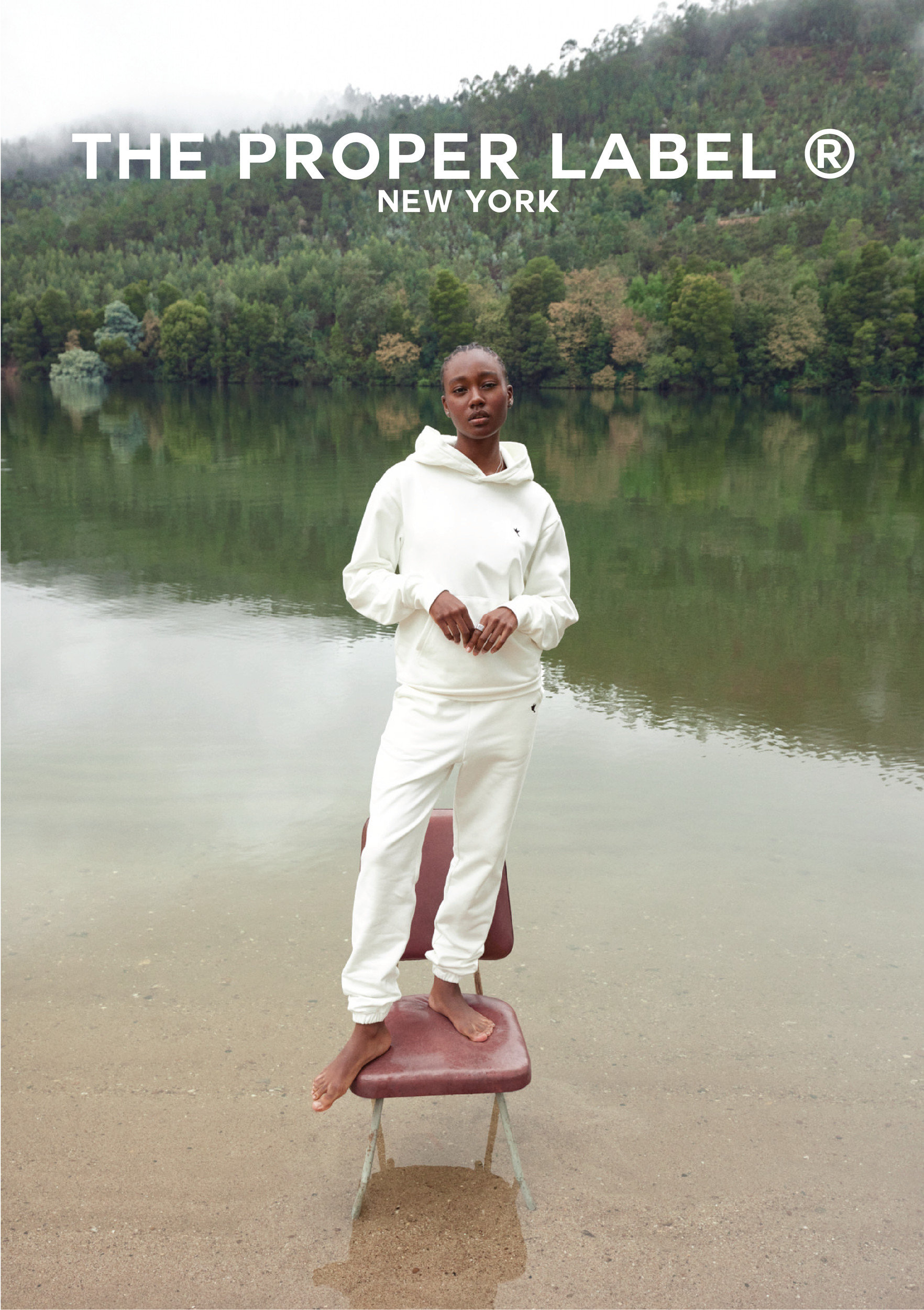 A model in white stands on a red chair at the edge of a lake.