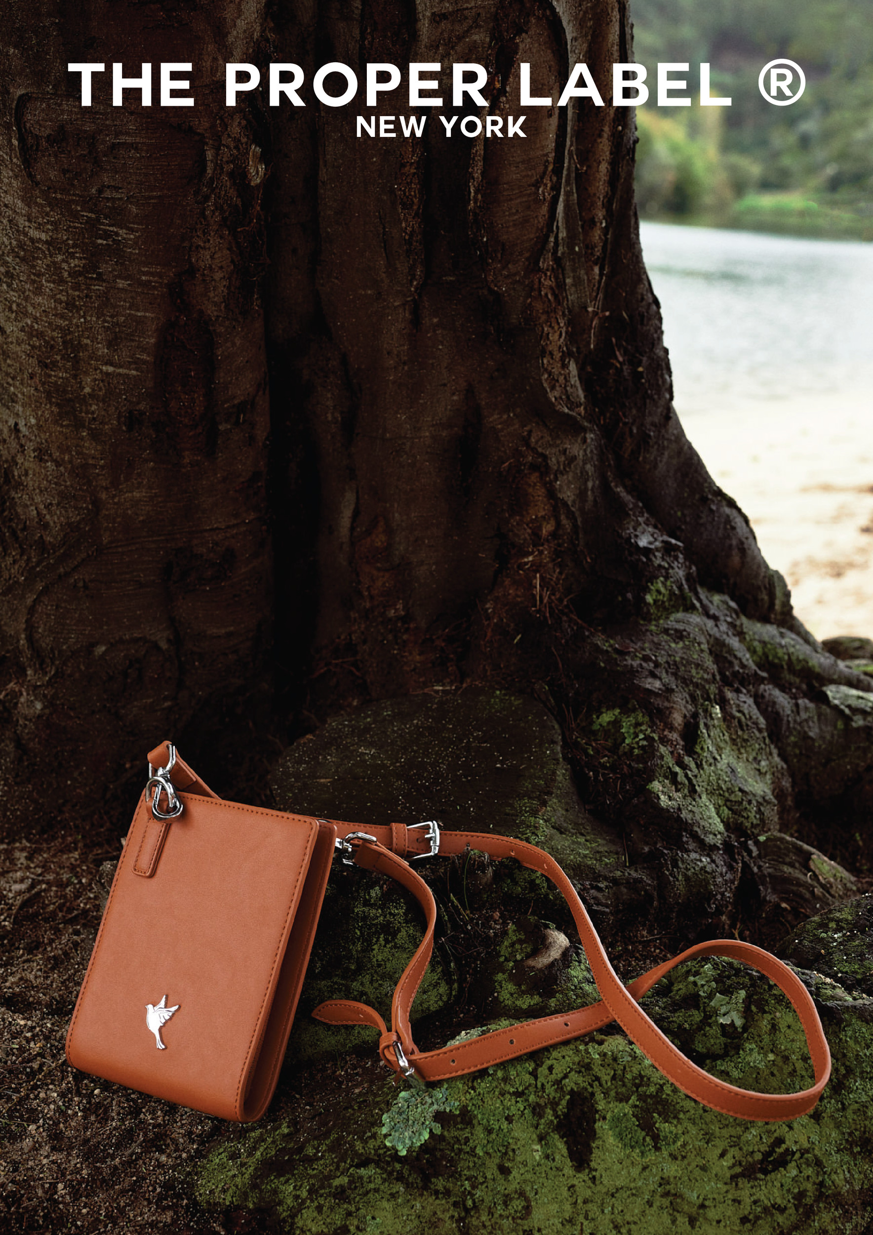 A brown leather crossbody bag rests against a tree.